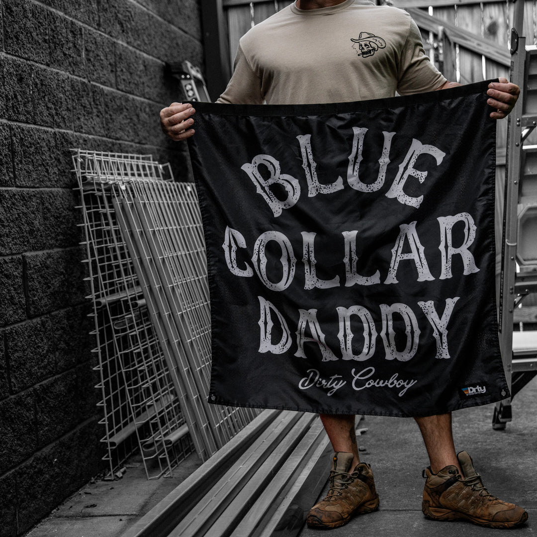 A person outdoors holding a black banner that reads 'BLUE COLLAR DADDY' with 'Dirty Cowboy' below in white lettering. The person is wearing a beige t-shirt, shorts, and brown boots, standing next to a dark brick wall with metal fencing.